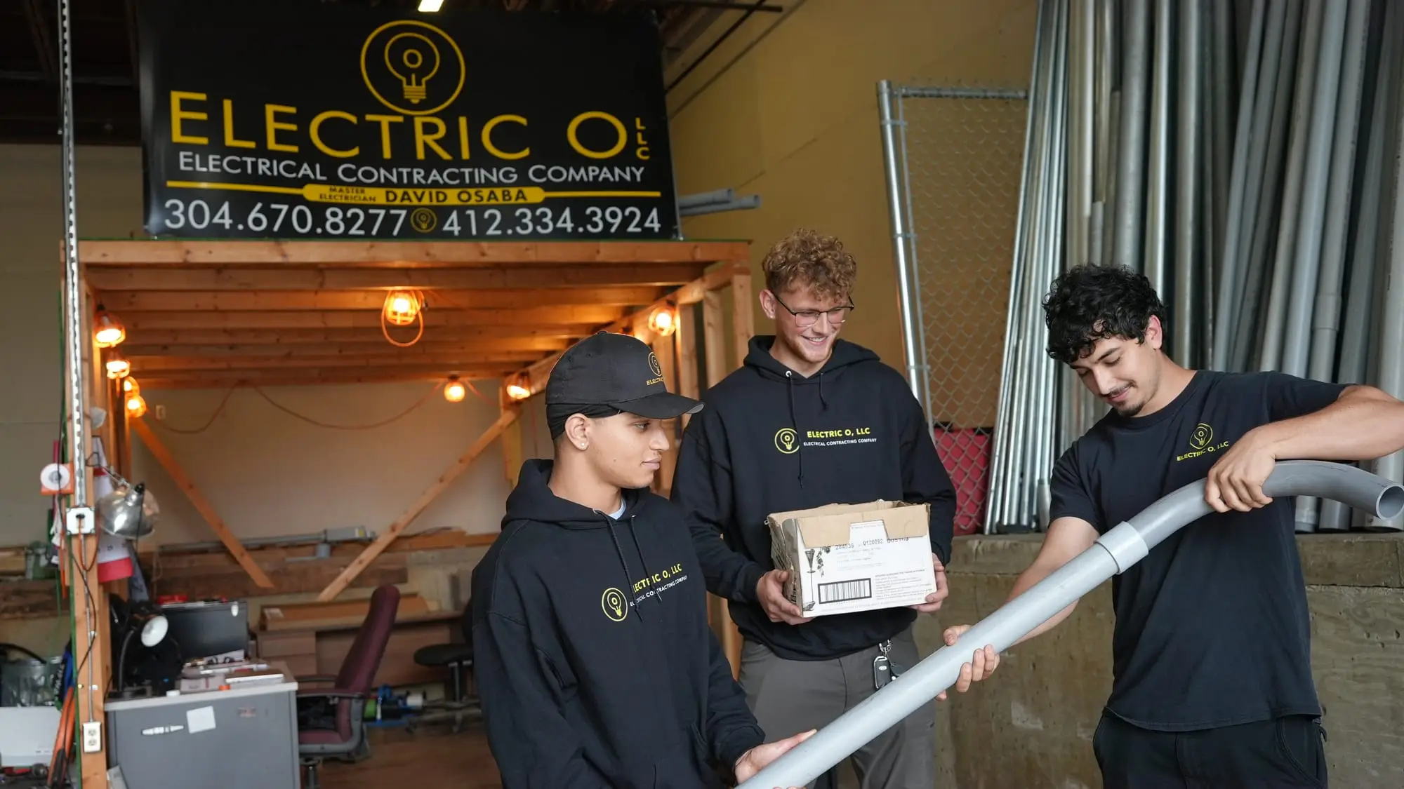 Three Electric O crew members in branded shirts and hoodies handling PVC conduit in the shop, conduit stock racked behind them.