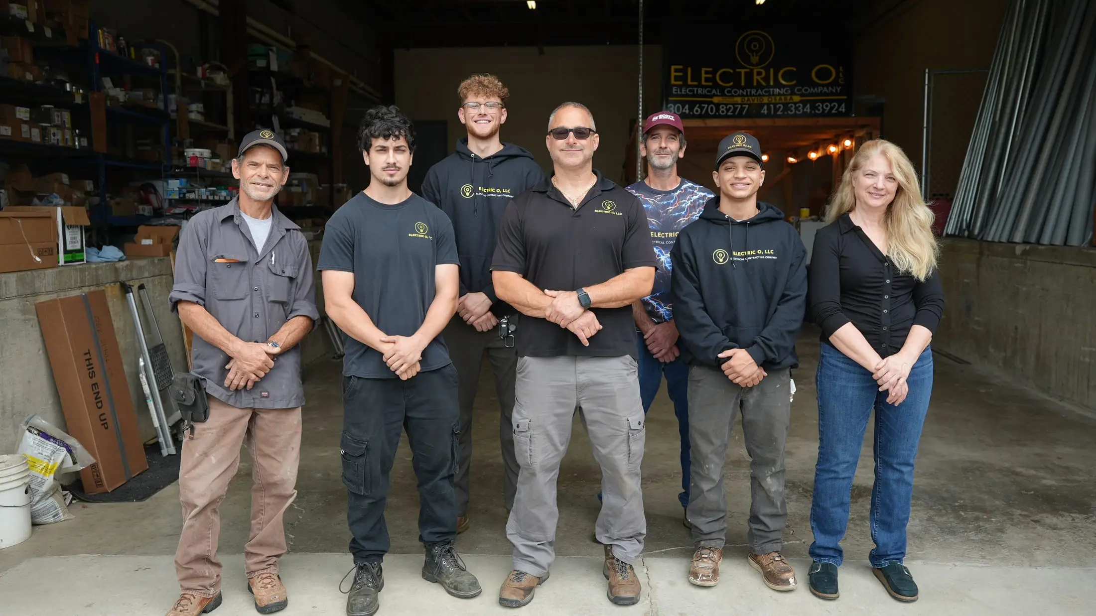 The seven people of Electric O in branded shirts and hoodies, standing in the shop under the Electric O banner with the company phone numbers.