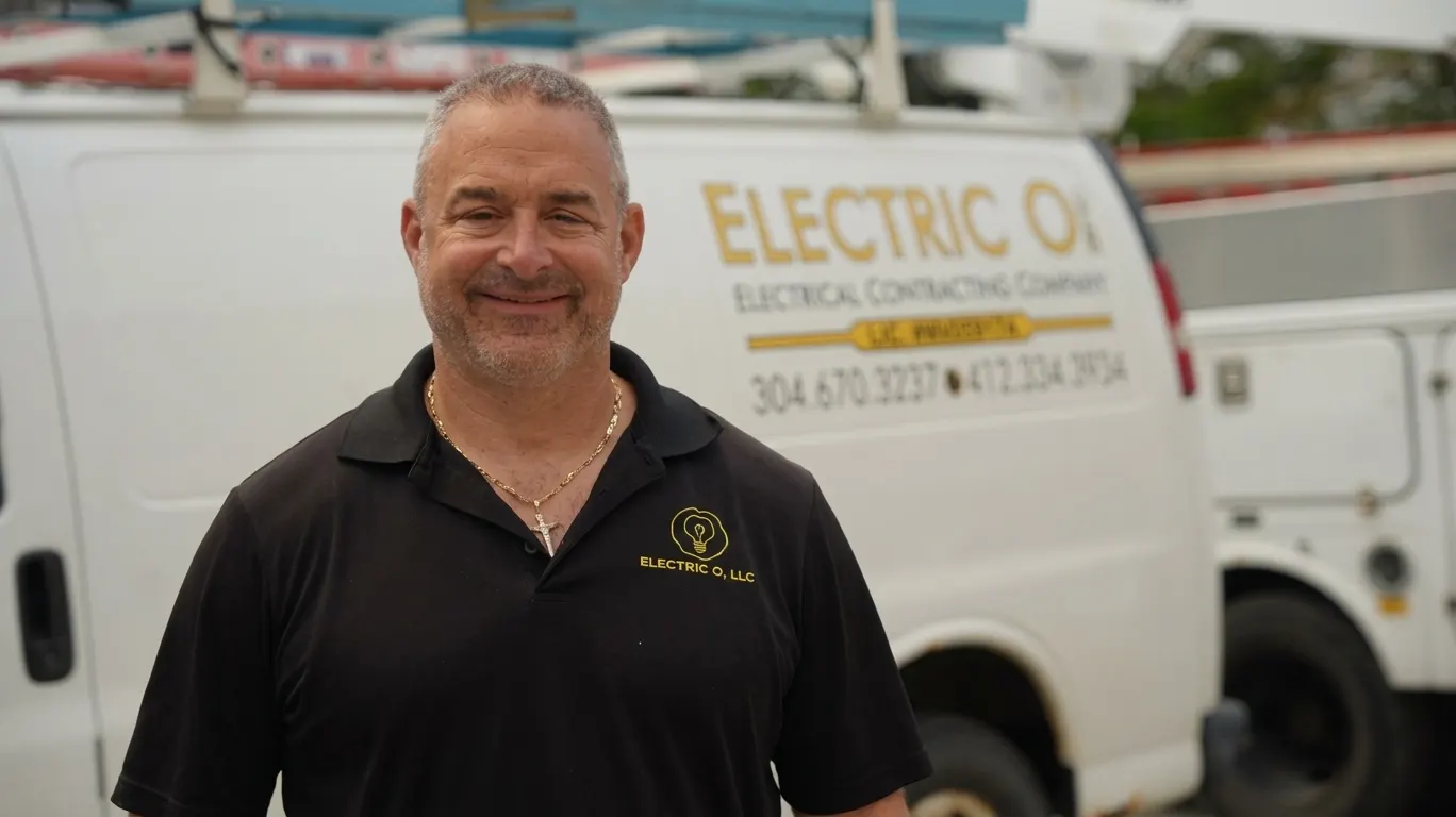 Dave Osaba smiling in a black Electric O polo in front of a white van lettered Electric O Electrical Contracting Company, with a second truck behind him.