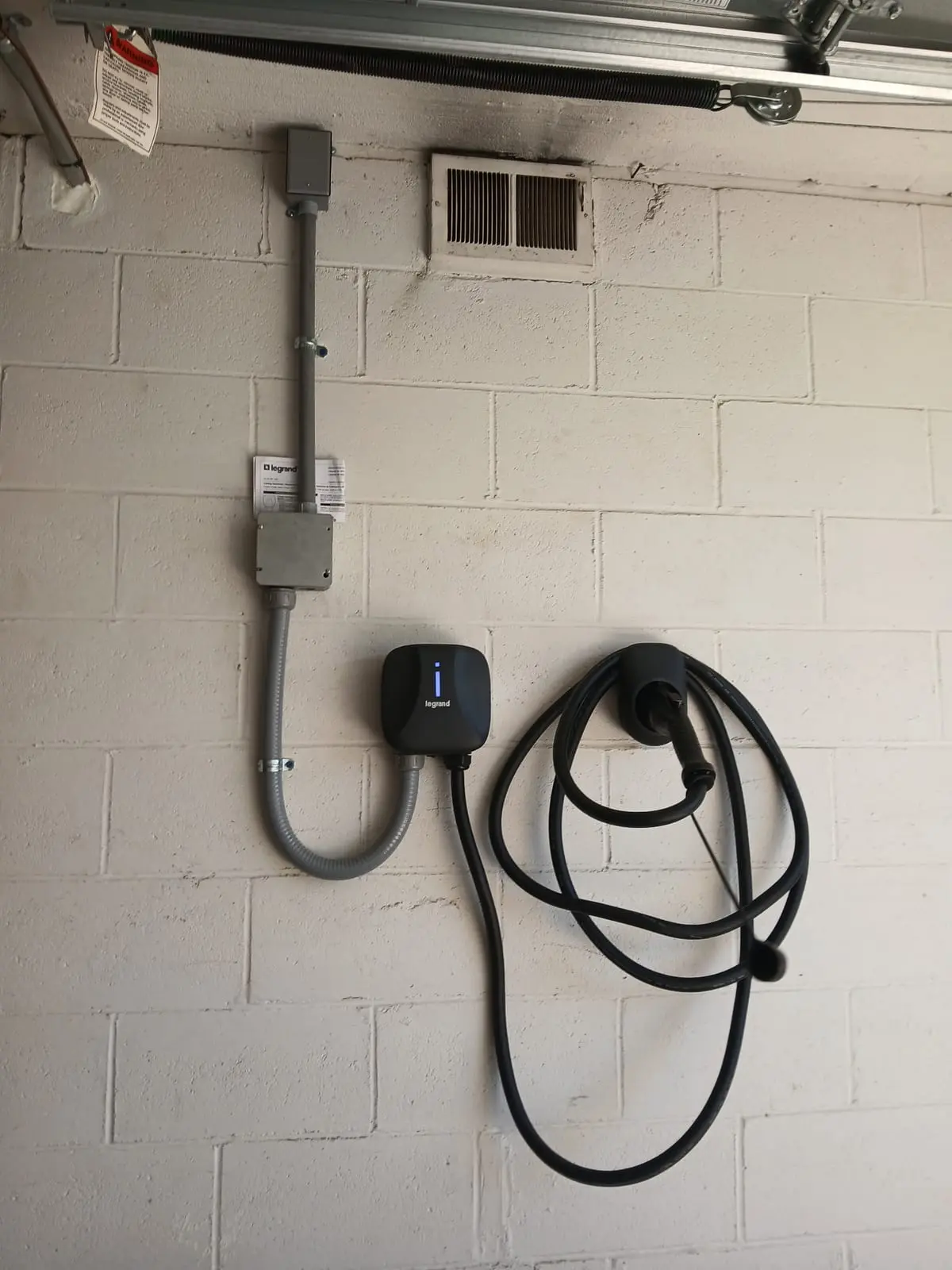 A Legrand Level 2 EV charger mounted on a painted block garage wall, with conduit running up from a junction box to the ceiling, installed by Electric O.