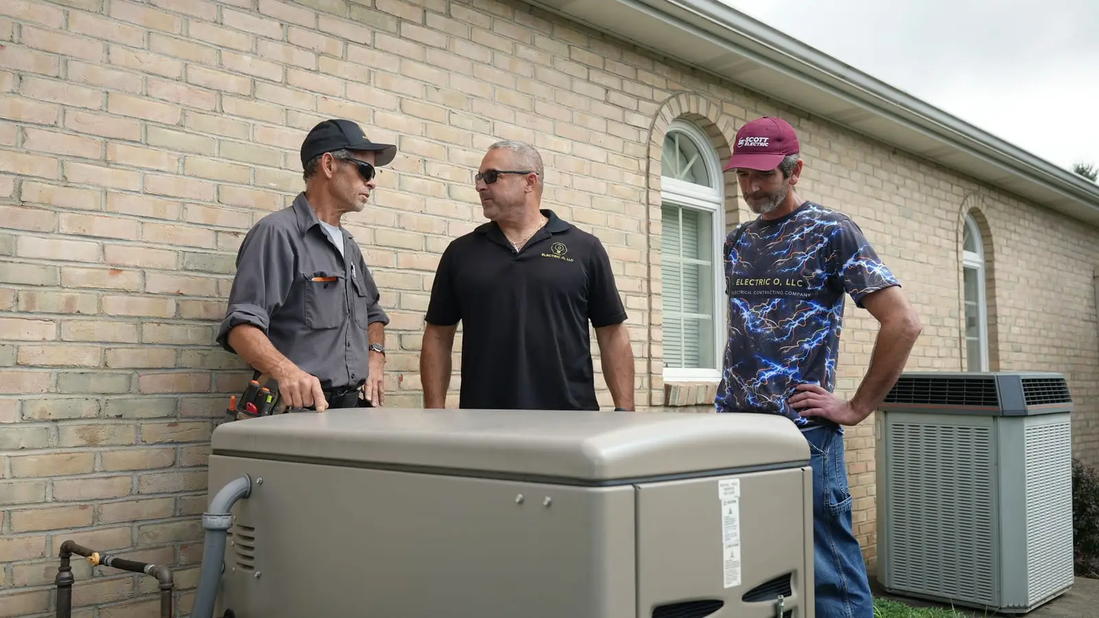 Three Electric O crew members standing behind a standby generator beside a tan brick house, talking over the install.