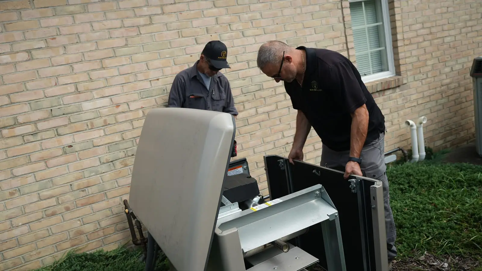 Two Electric O crew members working on a standby generator with its enclosure panels removed, beside a brick house.