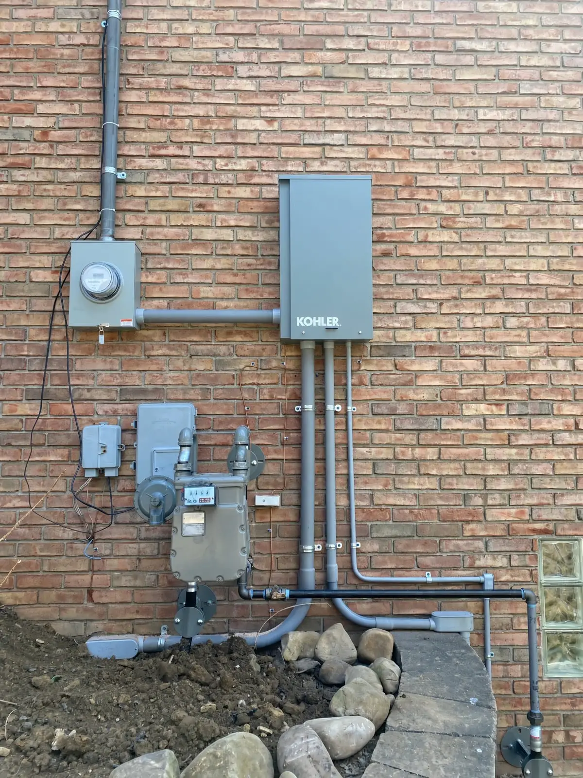 A Kohler automatic transfer switch mounted on a brick wall with conduit runs to the meter and gas line below.