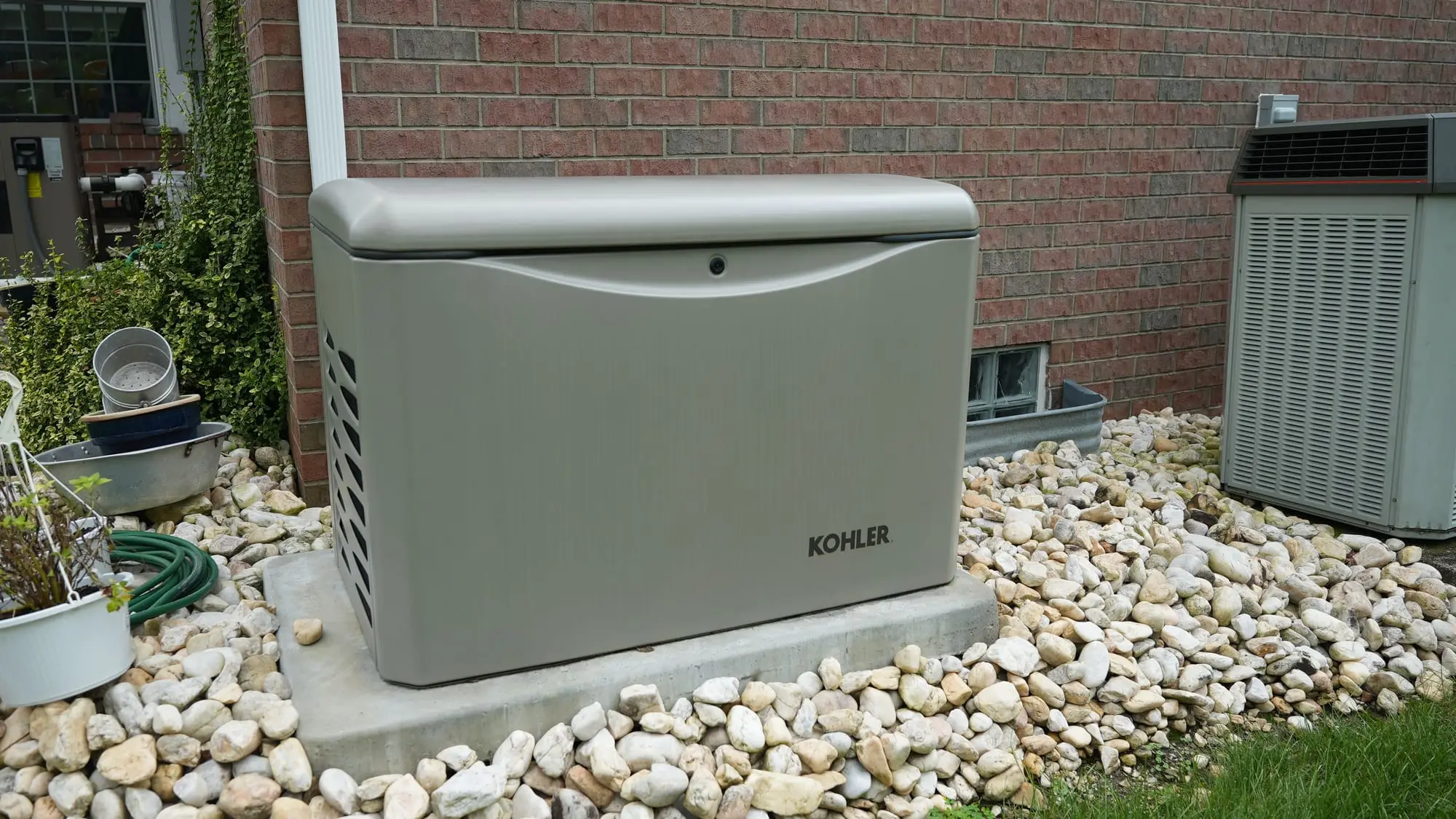 A Kohler standby generator on a concrete pad in a river-rock bed beside a brick house, next to the AC unit, installed by Electric O.