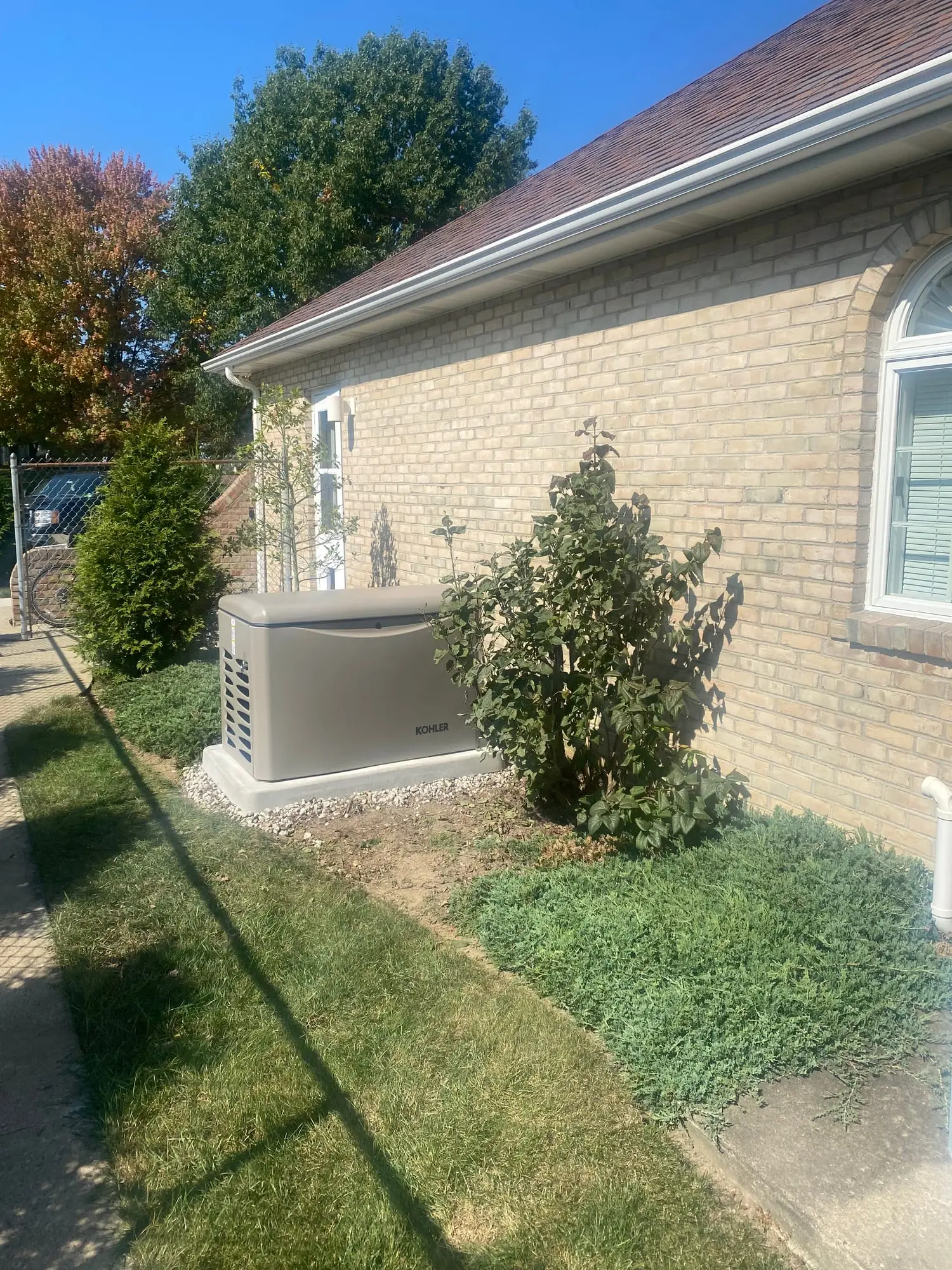 A Kohler standby generator on a gravel pad beside a brick home, next to a shrub and a fenced yard.