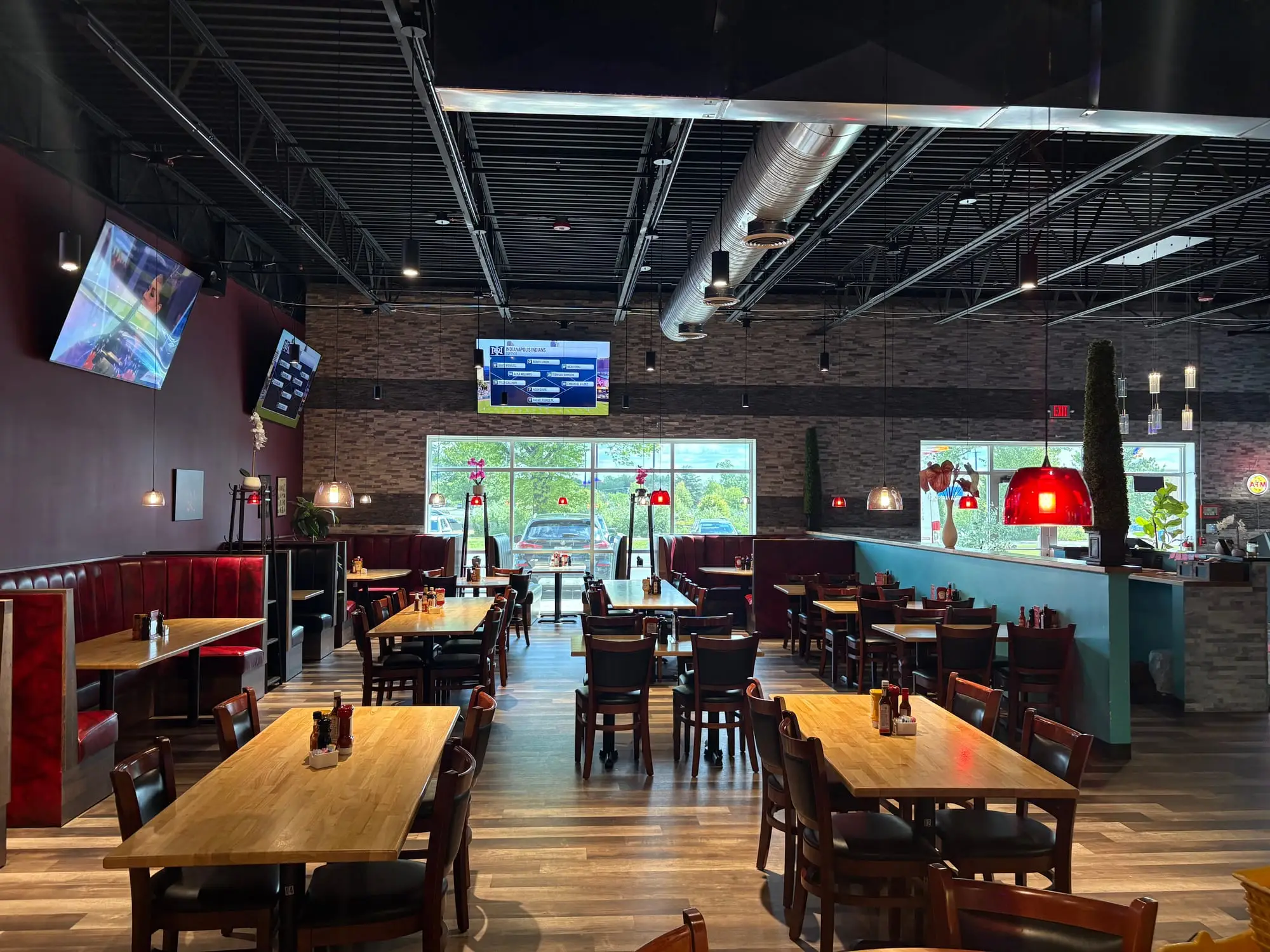 A restaurant dining room with red pendant lights over tables, booths, TVs and an exposed black ceiling.