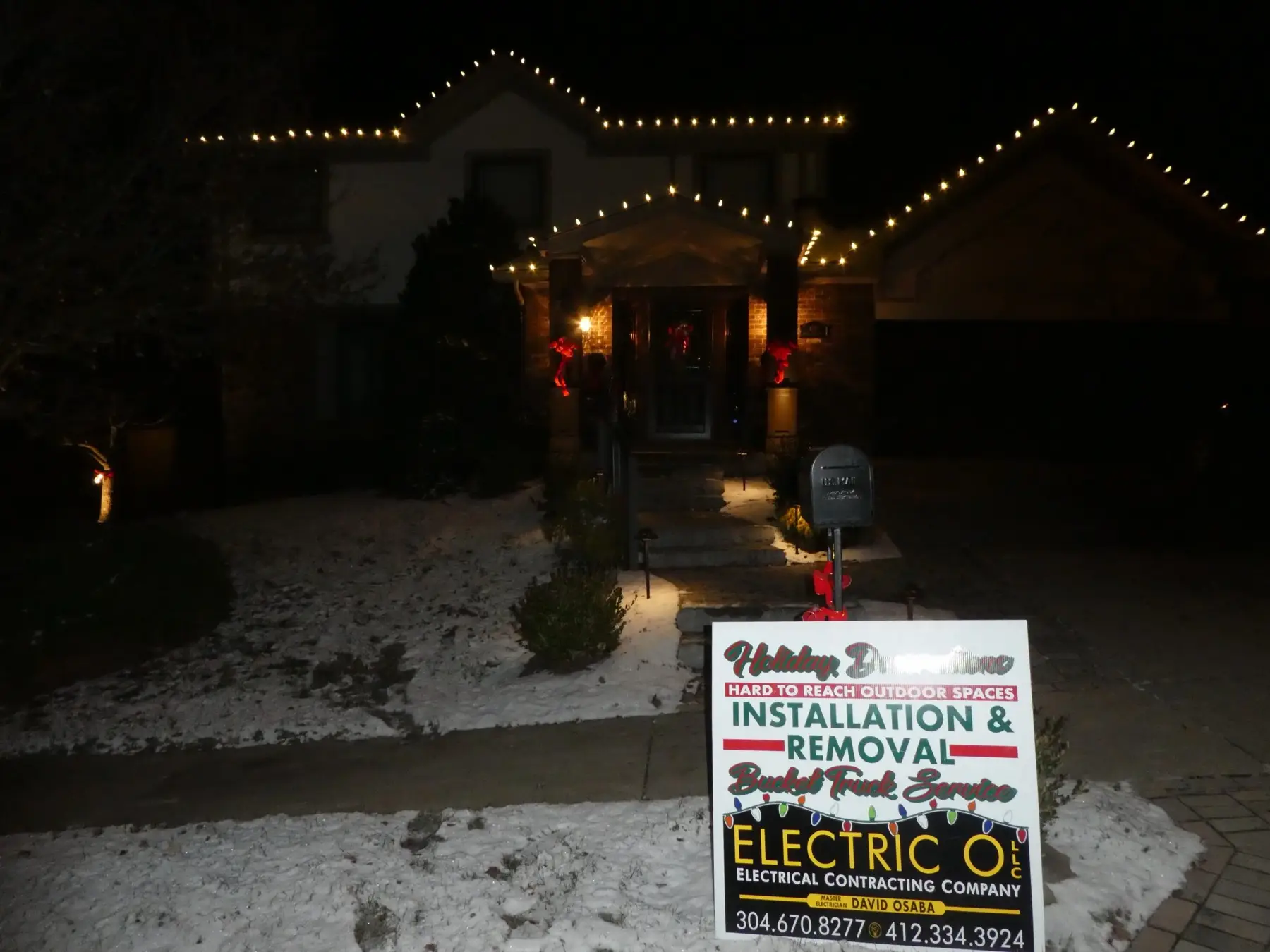 A house outlined in holiday lights at night with an Electric O yard sign in front.