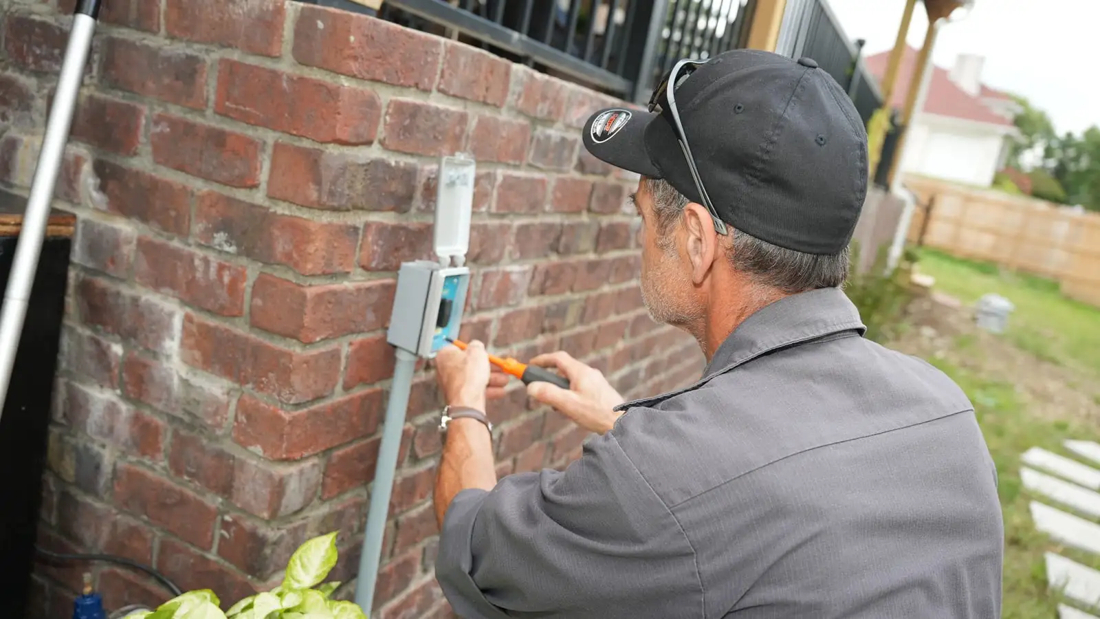 An Electric O electrician in a cap tightening a weatherproof outlet cover on a brick pillar, conduit running down to the ground.