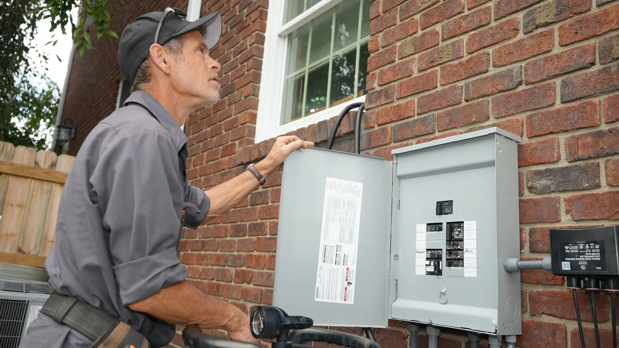 An Electric O electrician in a work shirt and cap inspecting an open outdoor breaker panel on a red brick wall.