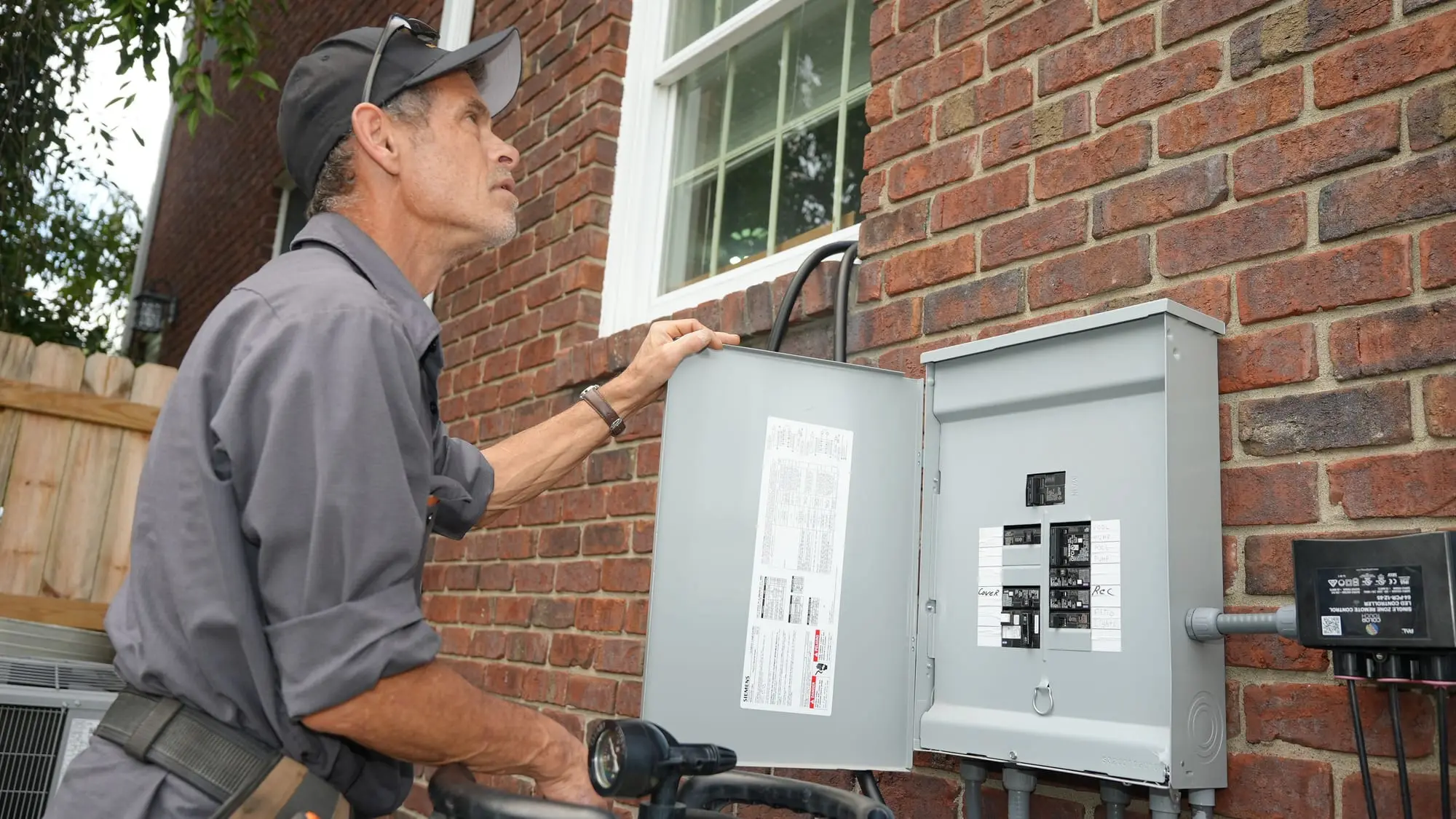 An Electric O electrician inspecting an open outdoor breaker panel on the brick wall of a home.