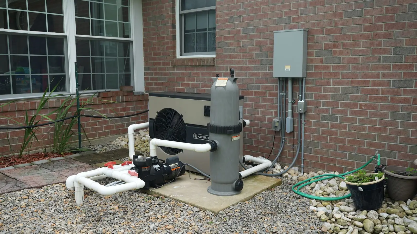 A pool heater, pump and filter against a brick house, wired in conduit to a gray subpanel and disconnects on the wall.