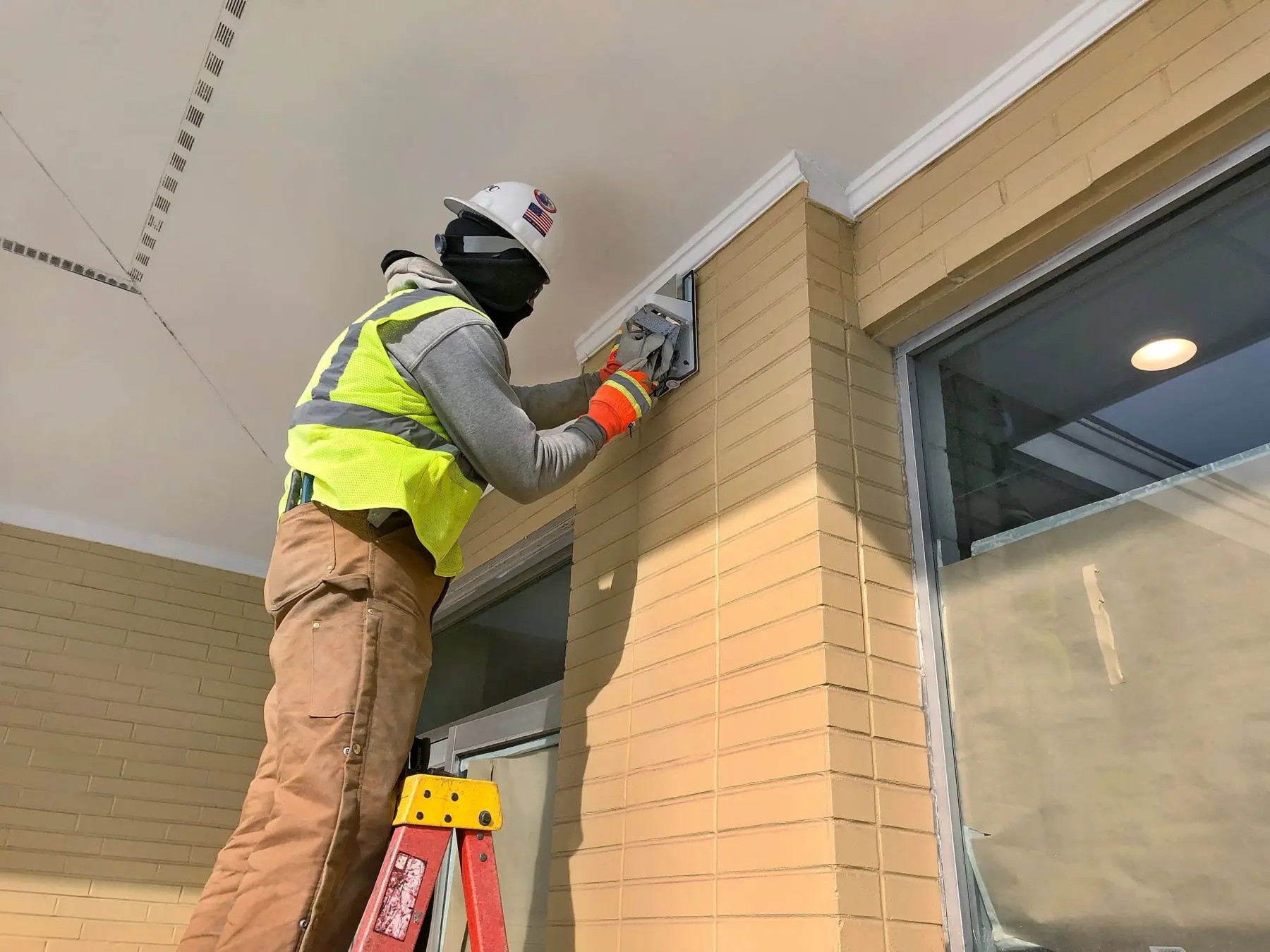 Electrician in a hard hat and hi-vis vest working on a soffit fixture from a ladder.