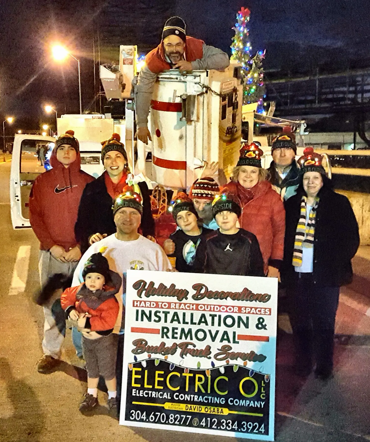 The Electric O crew and family posing with the bucket truck and a holiday sign at night.