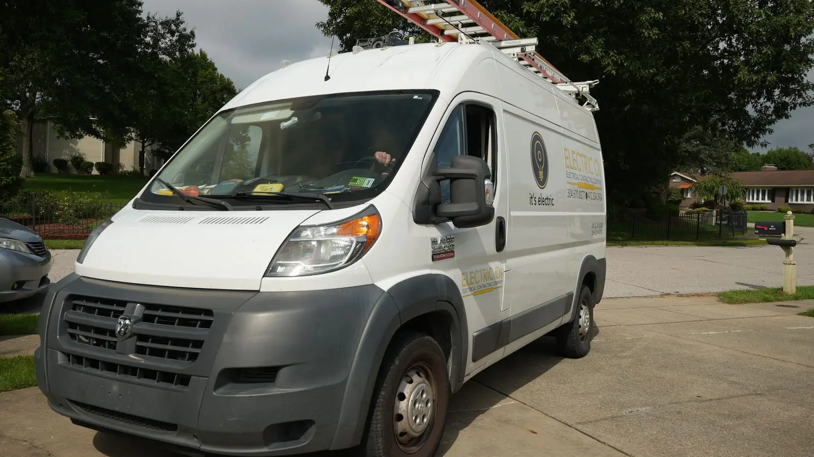 A white Electric O service van with ladders on its roof rack, parked in a residential driveway.