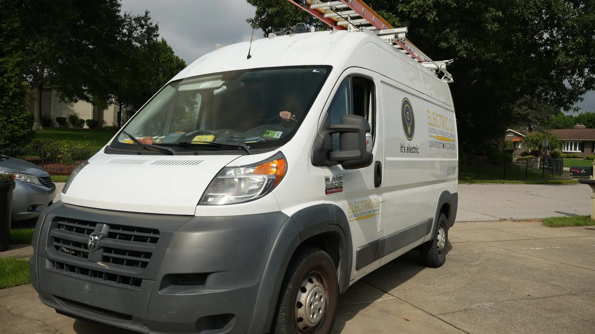 A white Electric O service van with ladders on the roof, parked in a residential driveway.