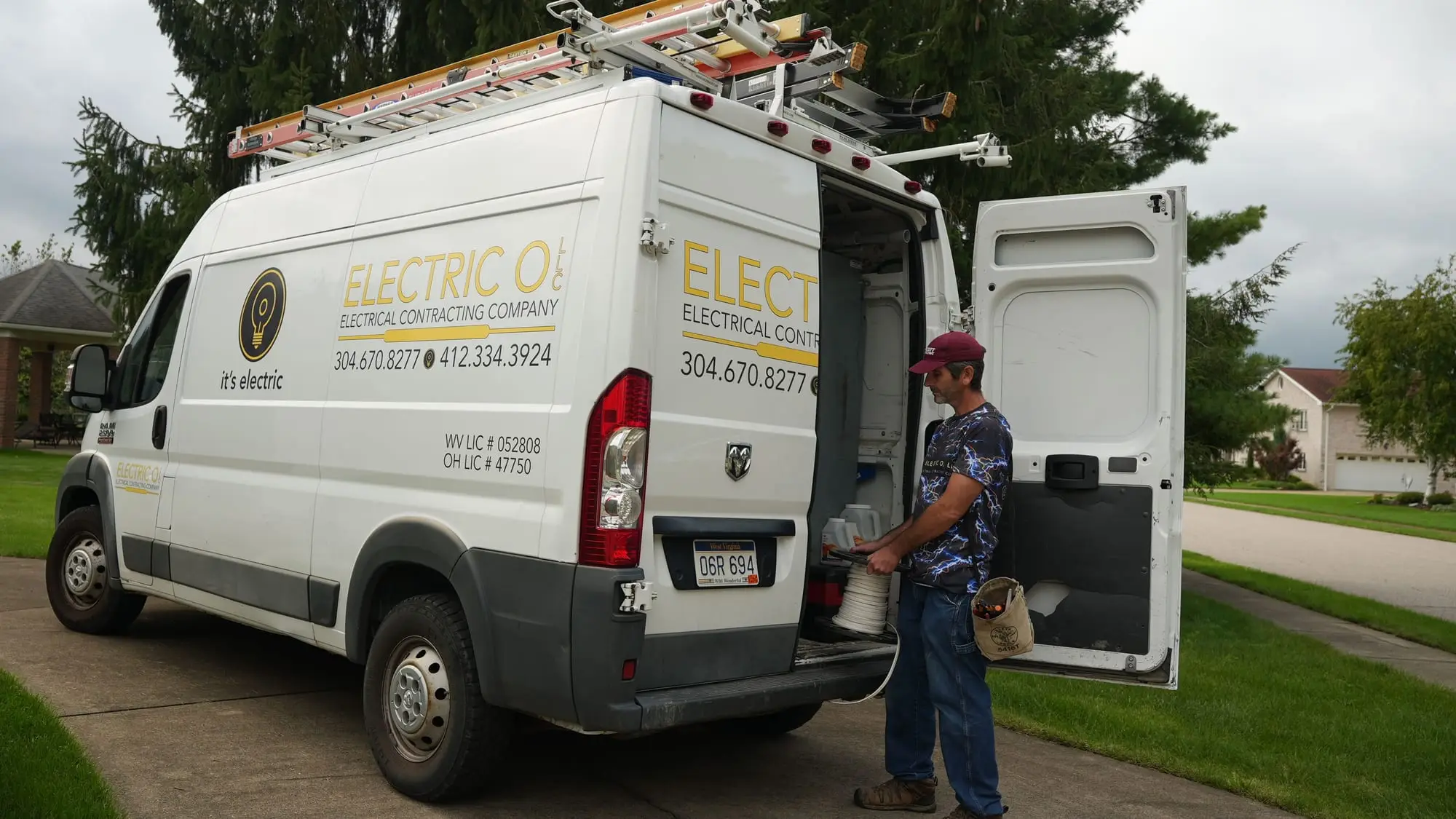 An Electric O service van with ladders on the roof and its back doors open in a residential driveway, a crew member unloading a spool of wire.