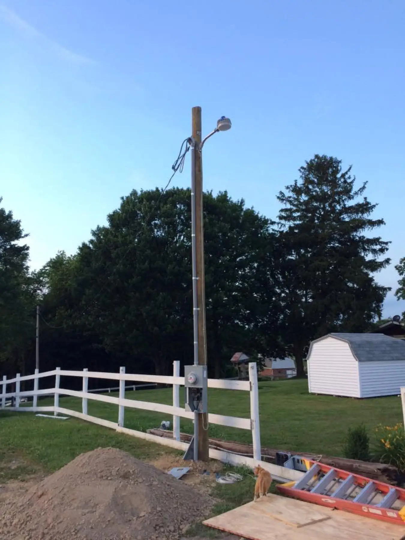 A new pole with a light and outlet box beside a white fence and shed.